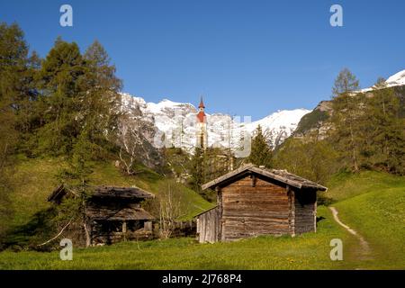 Alte Holzmühlen in Obernberg am Brenner, die historische Nikolaikirche im Hintergrund und Blumenwiesen im Vordergrund. Stockfoto