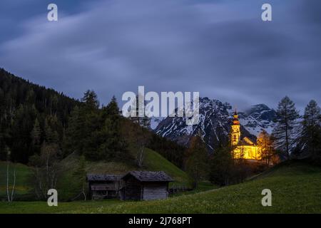 Alte Holzmühlen in Obernberg am Brenner, die historische Nikolaikirche im Hintergrund und Blumenwiesen im Vordergrund. Stockfoto