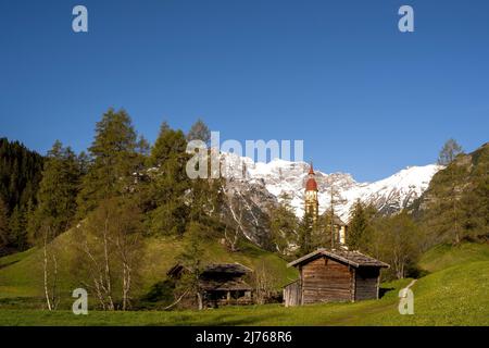 Alte Holzmühlen in Obernberg am Brenner, die historische Nikolaikirche im Hintergrund und Blumenwiesen im Vordergrund. Stockfoto