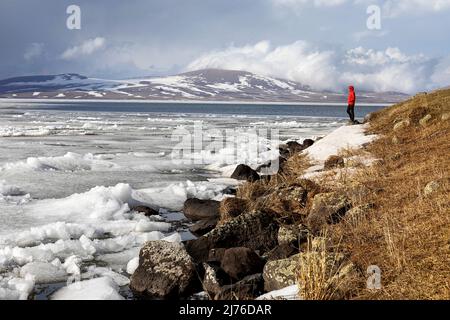 Frau in roter Jacke, die am Ufer des gefrorenen Paravani-Sees in Georgia steht, mit schneebedeckten Bergen im Hintergrund Stockfoto