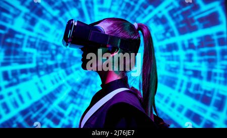 Seitenansicht einer Gamer-Frau mit Pferdeschwanz, der VR-Gerät trägt und vor blauem digitalem Hintergrund steht Stockfoto
