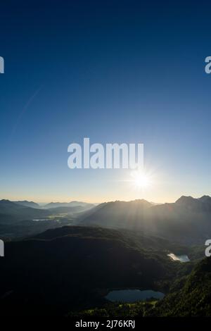 Sonnenstern über Karwendel, Ferchensee und Lautersee sowie über dem bewaldeten Kranzberg im Werdenfelser Land, an der Grenze zu Tirol/Österreich. Die beiden kleinen Seen liegen im dichten und dunklen Wald, während im Hintergrund die Sonne über dem Karwendel aufgeht. Stockfoto