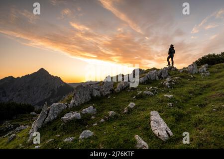 Bergsteiger auf einer Felsformation am Morgen rot auf dem Kompar im Karwendel. Im Hintergrund die Mondscheinspitze und eine tolle Morgenrot/Himmelsfarben. Der Gipfel der Mondscheinspitze sticht deutlich hervor, während die Silhouette des Mannes gegen den Himmel ragt. Stockfoto