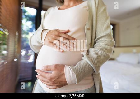 Mittlerer Abschnitt einer Schwangeren aus dem Kaukasus, die ihren Bauch berührt und zu Hause steht Stockfoto