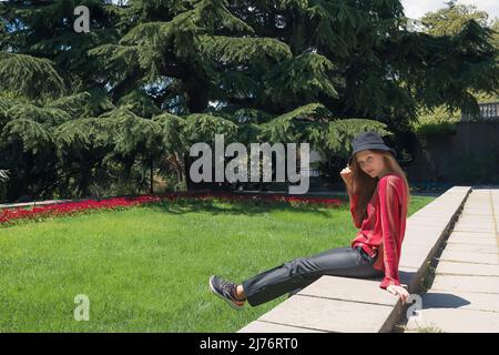 Hübsche junge Frau in einer roten Bluse und Hut läuft vor dem Hintergrund eines grünen Rasens aus Blumen und Fichte. Mädchen genießen Frühling Zeit. Frühlingsliebe Stockfoto