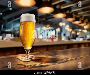 Gekühltes Glas Bier mit Kondensat auf dem Holztisch. Unscharfer Balken im Hintergrund. Stockfoto