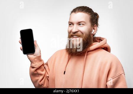 Skandinavischer gutaussehender glücklicher Mann, der das Telefon mit einem schwarzen Bildschirm-Modell hält und drahtlose Kopfhörer auf grauem Hintergrund isoliert hat. Kerl mit Ingwer-Hairsty Stockfoto