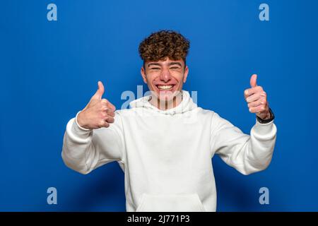 Attraktiver, fröhlicher junger Mann mit lockigen Haaren, der ein weißes Sweatshirt trägt, das isoliert auf blauem Hintergrund steht und die Daumen hoch zeigt, während er lächelt und Stockfoto