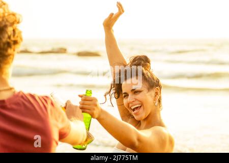 indische hispanische Frau im Fokus hält Flasche mit Limonade oder Bier und klickt mit Freunden Stockfoto