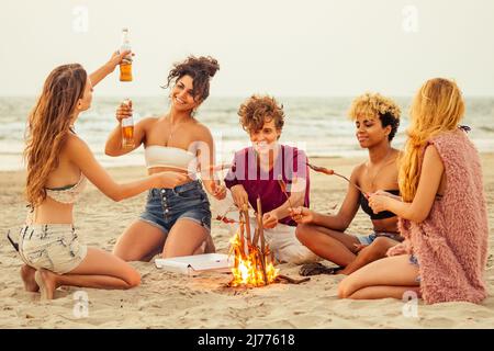 Gemischtes Rennen multinationale Gruppe von Freunden kochen Würstchen und trinken Limonade bei Sonnenuntergang auf Sand Stockfoto