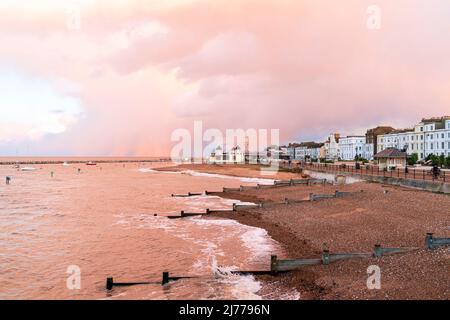 Herne Bay mit Wolken, die vom Sonnenuntergang erhellt werden (nicht gesehen). Bei Flut, mit Groynes am Strand, Uhrenturm und Regenwolken am Horizont. Stockfoto