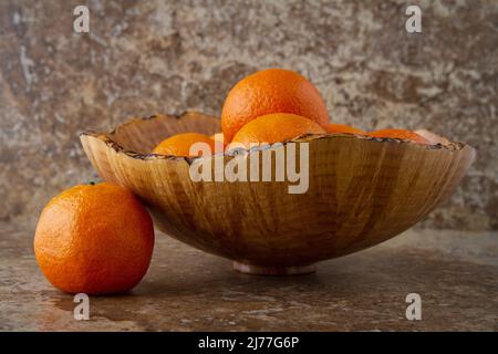 Ein Stillleben einer dekorativen Holzschale gefüllt mit Mandarinen auf warm getönten braunen Fliesen mit einer einzigen Frucht neben der Schale. Stockfoto