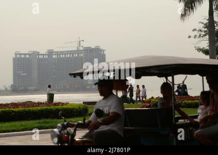 Menschen, die auf einem Fußgängerweg am Fluss gehen, im Vordergrund eines Tuk-Tuks, das Passagiere in Phnom Penh, Kambodscha, befördert. Stockfoto