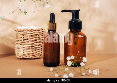 Kosmetische Gesichtsreiniger und braune Glasflaschen aus Serum, Ohrstöcke im Korb und weiße Gypsophila-Blüten auf hellbeigem Hintergrund. Naturkosmetik, Stockfoto