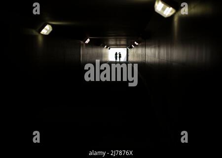 Zwei Silhouetten am Ende des Tunnels. Licht in der Ferne. Figuren von kleinen Frauen in kontrastierendem Licht. Lampen in langer Unterführung. Stockfoto