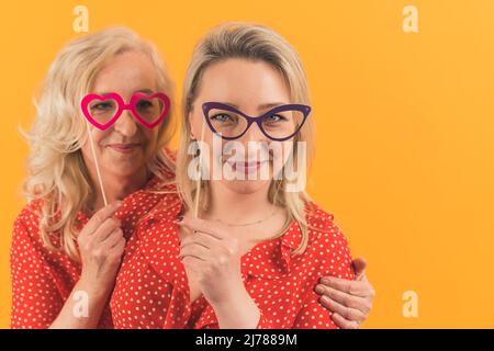 Zwei schöne Frauen, Mama mit Tochter, die Spaß zusammen haben, während sie auf lustige bis bunte Brille, Familienkonzept, isoliert, Copyspace, oranger Hintergrund. Hochwertige Fotos Stockfoto
