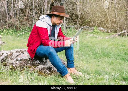 Selbstständiger Unternehmer, der Hut trägt, auf sein Tablet schaut, während er sich auf dem Feld entspannt und auf einem Stein sitzt. Konzept der Arbeit, genießen, entspannen, ich Stockfoto