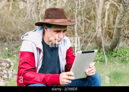 Selbstständiger Unternehmer, der Hut trägt, auf sein Tablet schaut, während er sich auf dem Feld entspannt und auf einem Stein sitzt. Konzept der Arbeit, genießen, entspannen, ich Stockfoto