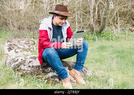 Selbstständiger Unternehmer, der Hut trägt, auf sein Tablet schaut, während er sich auf dem Feld entspannt und auf einem Stein sitzt. Konzept der Arbeit, genießen, entspannen, ich Stockfoto