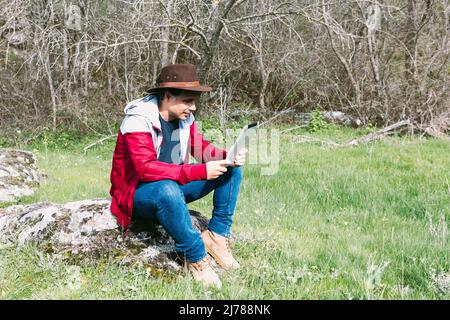 Selbstständiger Unternehmer, der Hut trägt, auf sein Tablet schaut, während er sich auf dem Feld entspannt und auf einem Stein sitzt. Konzept der Arbeit, genießen, entspannen, ich Stockfoto