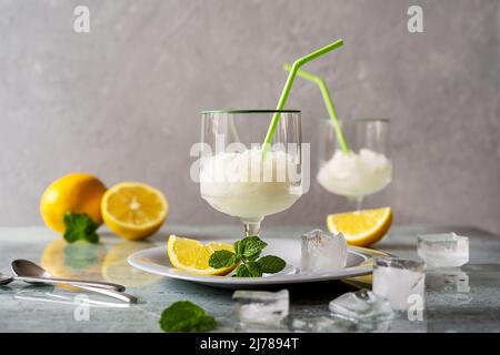 Zitronensorbet in zwei Gläsern auf Teller mit Zitronen, Eiswürfeln, Minzblättern und Trinkhalmen auf grauem Hintergrund Stockfoto