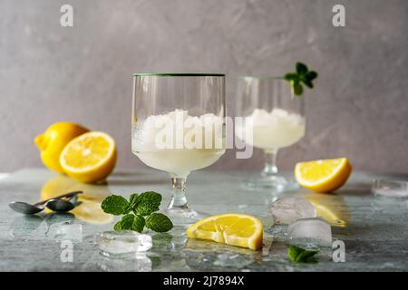 Zitronensorbet in zwei Gläsern mit Zitronen, Eiswürfeln, Minzblättern auf grauem Hintergrund Stockfoto