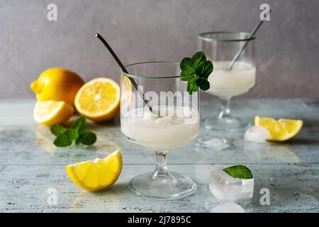 Zitronensorbet in zwei Gläsern mit Zitronen, Eiswürfeln, Teelöffeln, Minzblättern auf grauem Hintergrund Stockfoto