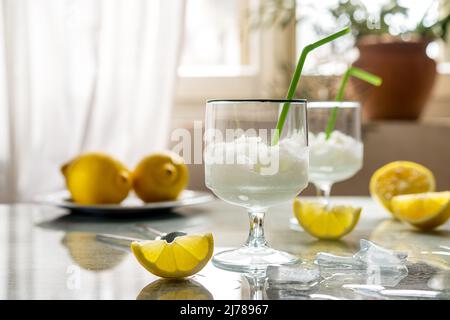 Zitronensorbet in zwei Gläsern mit Zitronen, Eiswürfeln und Trinkhalmen auf grauem Hintergrund und Fensterrückseite Stockfoto
