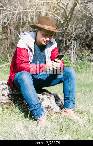 Selbstständiger Unternehmer mit Hut, der auf sein Handy schaut, während er sich auf dem Land entspannt und auf einem Stein sitzt. Konzept der Arbeit, genießen, re Stockfoto