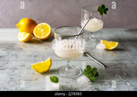 Zitronensorbet in zwei Gläsern mit Zitronen, Eiswürfeln, Teelöffeln, Minzblättern auf grauem Hintergrund Stockfoto