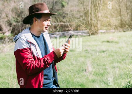 Unternehmerischer, selbstständiger Mann mit Hut, der auf sein Mobiltelefon blickt, während er sich auf dem Feld auf einer Wiese entspannt. Konzept der Arbeit, genießen, entspannen, Stockfoto