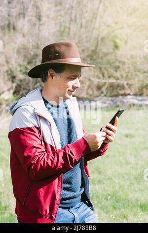 Unternehmerischer, selbstständiger Mann mit Hut, der auf sein Mobiltelefon blickt, während er sich auf dem Feld auf einer Wiese entspannt. Konzept der Arbeit, genießen, entspannen, Stockfoto