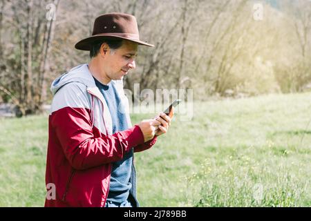 Unternehmerischer, selbstständiger Mann mit Hut, der auf sein Mobiltelefon blickt, während er sich auf dem Feld auf einer Wiese entspannt. Konzept der Arbeit, genießen, entspannen, Stockfoto