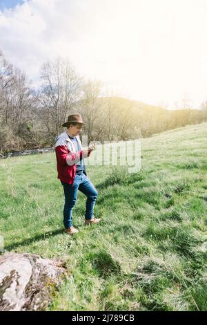 Unternehmerischer, selbstständiger Mann mit Hut, der auf sein Mobiltelefon blickt, während er sich auf dem Feld auf einer Wiese entspannt. Konzept der Arbeit, genießen, entspannen, Stockfoto