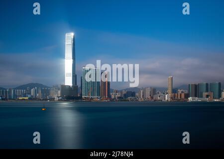 Die Sonne spiegelt sich in einem Wolkenkratzer auf Kowloon, Hongkong, und wirft Licht über das blaue Meer. Lange Exposition gibt ein seidig glattes Meer. Stockfoto