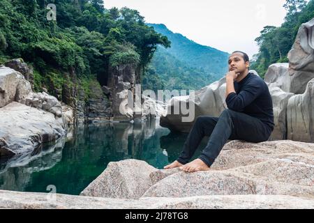 Isolierter junger Mann, der auf einem natürlich geformten weißen glänzenden Stein in einzigartiger Form am trockenen Flussbett sitzt, wird bei Sliang wah Umngot amkoi jaintia hi aufgenommen Stockfoto