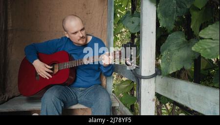 Der Mensch leitet eine Online-Akustikgitarre-Lektion von der Veranda seiner Vorstadtfarm aus Stockfoto