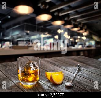 Ein Glas Whiskey mit Eiswürfeln im Inneren und zwei orangefarbenen Scheiben befinden sich auf dem alten Holztisch. Verschwommenes Interieur des Pubs im Hintergrund. Stockfoto