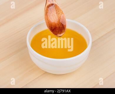 Weiße Schale mit Sonnenblumenöl auf Holzgrund. Öl aus einem Holzlöffel fließt in eine Schüssel. Stockfoto