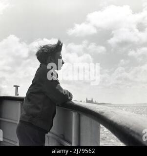 1970, historisch, ein Junge auf einem Boot, auf der Holzschiene angelehnt, mit Blick auf den Fluss Mersey, Liverpool, England, Großbritannien. Das Royal Liver Building ist in weiter Ferne zu sehen. Stockfoto