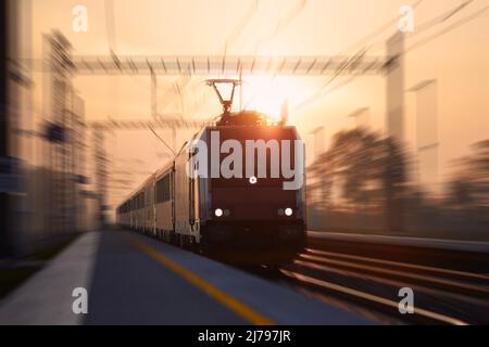 Der Personenzug fährt schnell durch den Bahnhof. Bahn bei Sonnenaufgang mit Unschärfe-Effekt. Stockfoto