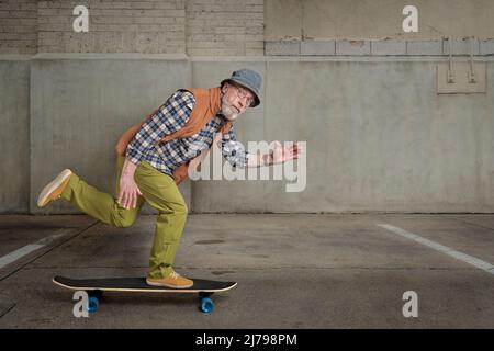 Bärtiger älterer Mann mit runder Brille und einem Eimer Hut reitet auf einem langen Skateboard in einer grunge städtischen Umgebung Stockfoto