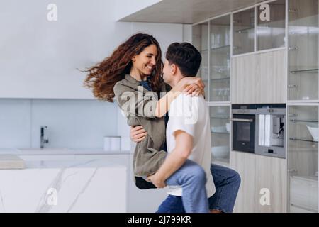 Der junge Brunet-Mann hält sich in den Armen fest und spinnt die lockige Frau in Hemd und Jeans, umarmt sich fest und lacht in der Küche zum Lachen Stockfoto