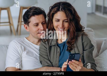 Der junge rothaarige Ehemann und die lockige Frau diskutieren über Hochzeitsfotos aus den sozialen Medien, lächeln breit und sitzen in der Nähe auf dem Sofa Stockfoto