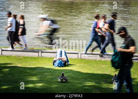 07. Mai 2022, Hessen, Frankfurt/Main: Ein Mann ruht sich in der Sonne auf dem Rasen am Mainufer in Frankfurt aus, während Menschen um ihn herum laufen. Foto: Frank Rumpenhorst/dpa Stockfoto