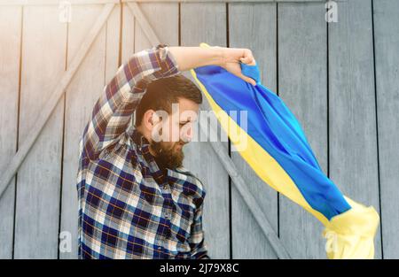 Der Mann in einem karierten Hemd hält bei schneebedecktem und sonnigem Wetter die Staatsflagge der Ukraine im Freien. Ukrainische Flagge, Patriotismus. Stockfoto