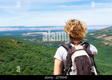 Die blonde Frau mit lockigen Haaren auf dem Rücken, mit einem Rucksack auf dem Rücken, auf einem Hügel, der in ein Tal mit einem Buchen- und Kiefernwald auf dem Hügel blickt Stockfoto