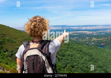 Die blonde, lockige Frau auf dem Rücken, mit einem Rucksack auf dem Rücken, auf einem Berg, zeigt mit der Hand auf ein Tal mit einer Buche und einer Kiefer Stockfoto