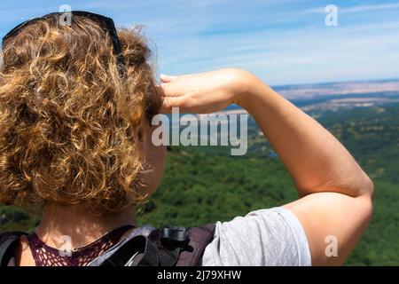 Blonde Frau auf dem Rücken mit Rucksack, auf einem Berg mit der Hand auf der Stirn und Blick auf den Horizont zu einem Tal mit Buche und Nadel Stockfoto