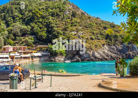 Ein junges Paar, das auf einem Motorradpark in der Nähe einer kleinen Bucht am Strand von Paleokastritsa reitet, mit Geschäften und Cafés auf der Insel Korfu, Griechenland. Stockfoto